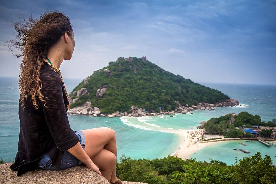 A girl is sitting on a mountain while traveling alone. Mountains and sea are visible in the distance